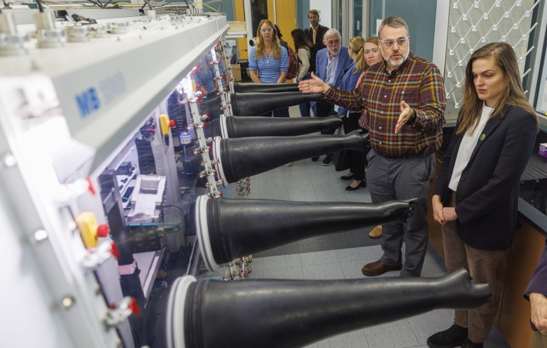 A man in a plaid shirt and safety glasses gestures toward a long glovebox with black gloves extending into the room, explaining it to a woman in a dark blazer, as a group of visitors looks on behind them.