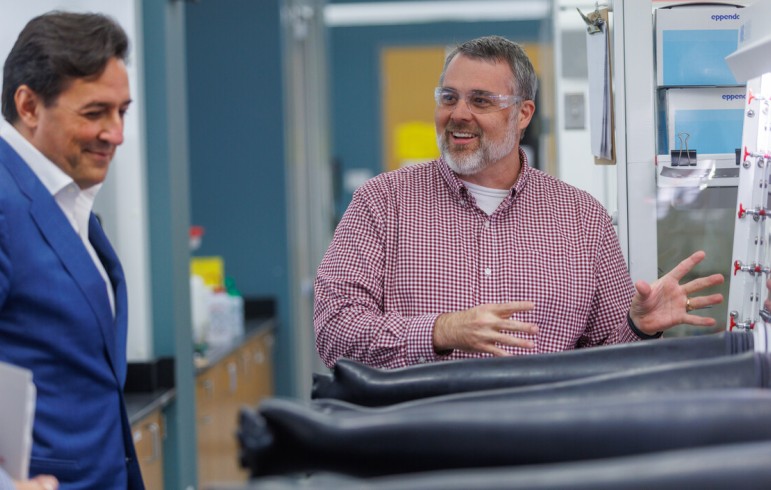 A man in a blue blazer looks on with a smile as a bearded man in a red checked shirt and safety glasses gestures animatedly beside a glovebox with black gloves in a laboratory.