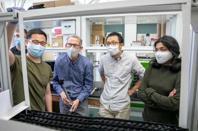 Four people in masks look into a rectangular metal frame in a lab