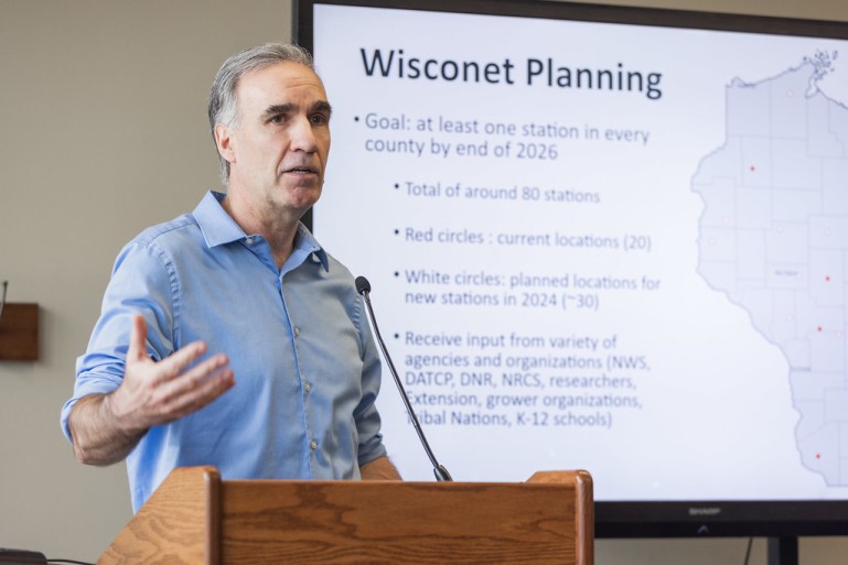 Man in blue shirt stands behind a podium gesturing with one hand. A screen behind him is titled "Wisconet Planning"