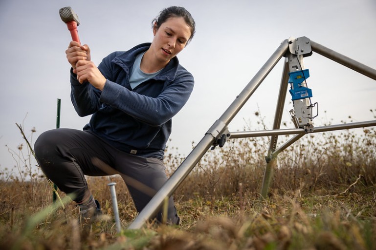 A woman swings a hammer towards a stake attached to a metal tripod sitting in a grassy field