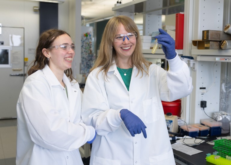 A woman in a white lab coat and safety glasses holds up a flask of opaque yellow liquid as another woman in similar attire looks on smiling