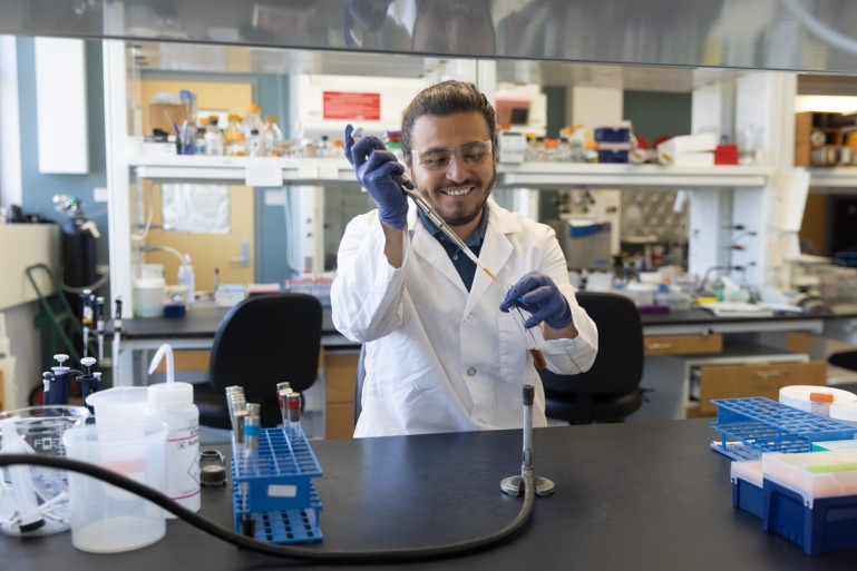 A smiling scientist in a white lab coat and purple nitrile gloves works with a pipette while standing at a black lab bench surrounded by various laboratory equipment including test tube racks, bottles, and other research instruments. 