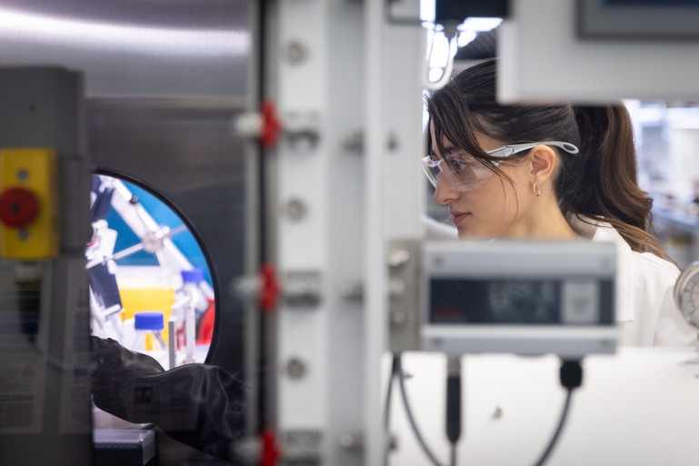 A researcher in a white lab coat and safety goggles looks through a circular portal into an automated laboratory workstation filled with robotic equipment and chemical vials.