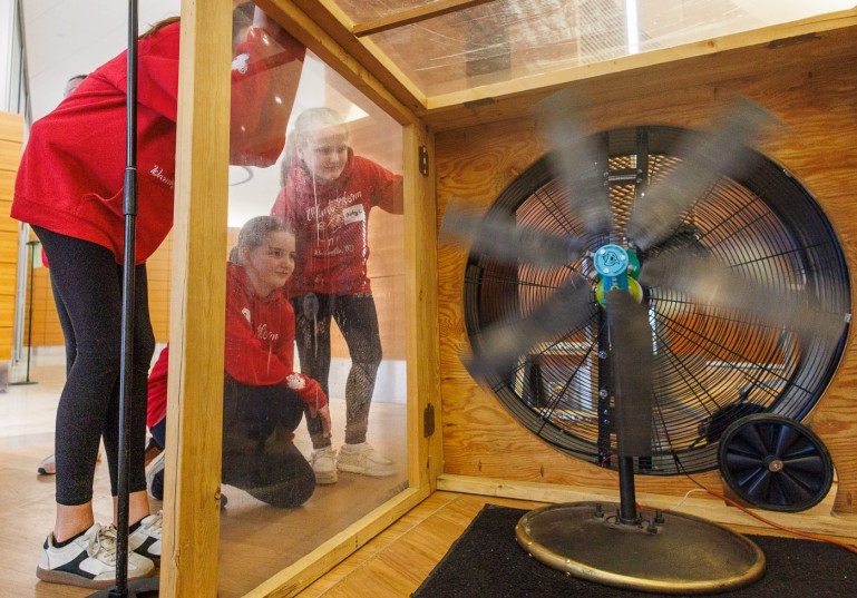 Three girls wearing matching red "Wind Storm" hoodies look through a clear panel into a wooden wind tunnel, watching a large fan spin rapidly.