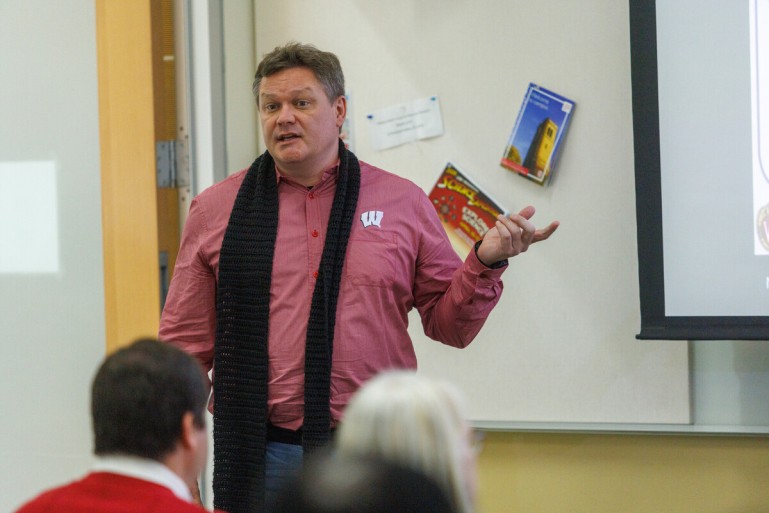 Man in red shirt with black scarf gestures as he talks to a room full of people