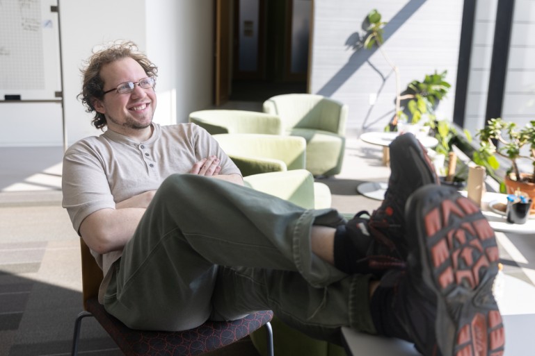A man reclines in a chair with his feet on a table in a sunlit space with potted plants