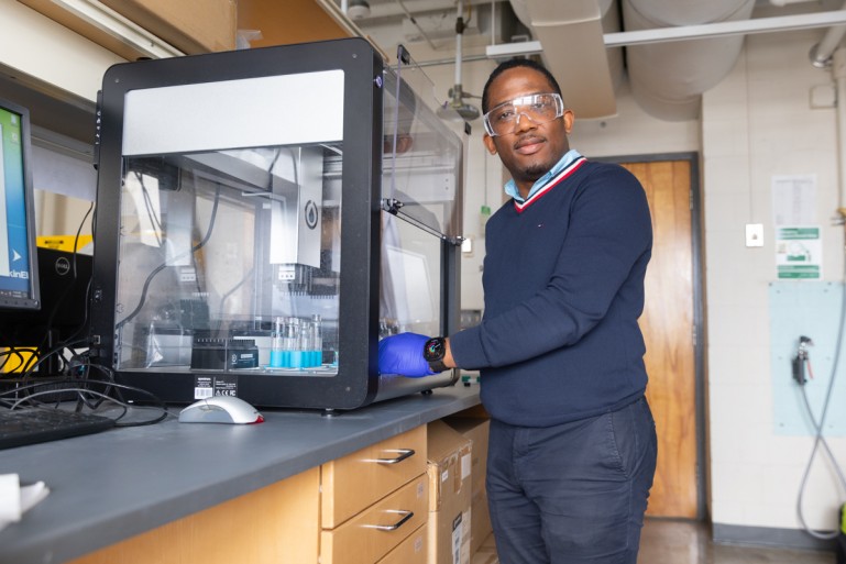 Man standing at a lab bench in front of a machine encased in metal and glass