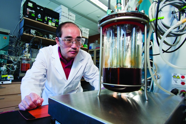 Man in white lab coat and protective glasses peers at flask of brown liquid in lab