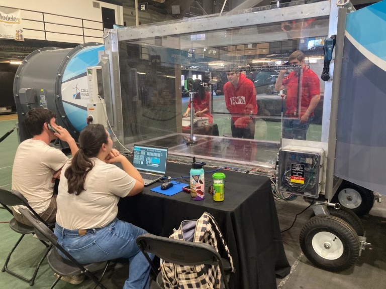 Two UW-Madison students sit at a control station with laptops, observing a model wind turbine being tested inside a clear-walled wind tunnel. Three people in red Wisconsin shirts work behind the transparent enclosure