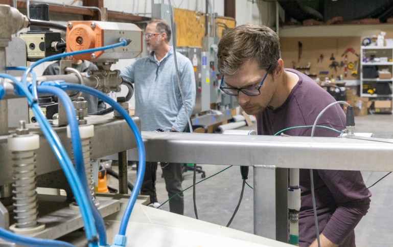 A man peers down at a piece of machinery with coil springs and blue tubingin an industrial setting. In the background a second man looks at something off camera. 