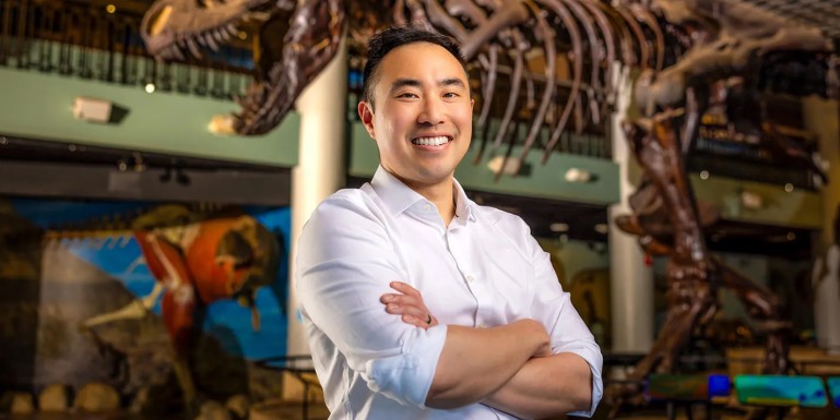 A smiling man with short dark hair, wearing a white button-down shirt with sleeves rolled up, stands with arms crossed in front of a large dinosaur skeleton display in a museum exhibit hall.