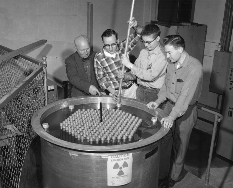 Four men stand over a cylindrical tank labeled with radiation warning. An older man on the left points towards the surface of water in the tank as two younger men hold a rod extending below the surface.