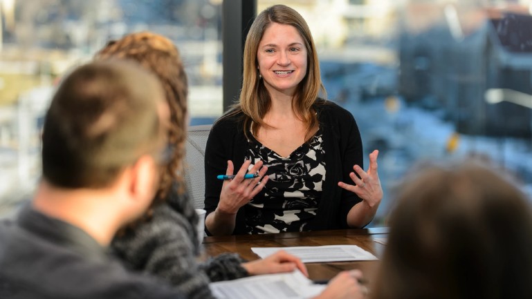 a woman smiles and gestures while speaking at a table with colleagues, whose backs are visible in the foreground, papers spread on the table in front of them, with a large window in the background.