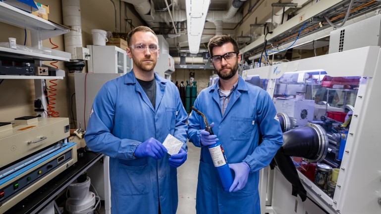 Two men in blue lab coats, gloves, and safety glasses pose holding objects in a laboratory