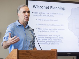 Man in blue shirt stands behind a podium gesturing with one hand. A screen behind him is titled "Wisconet Planning"