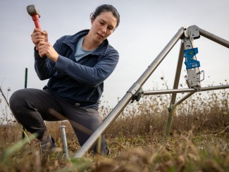 A researcher hammers a ground stake to install field monitoring equipment on a tripod in a grassland or prairie field under an overcast sky.