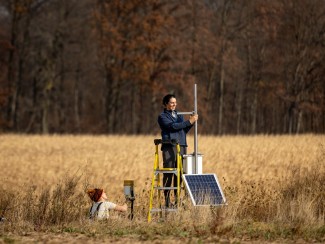 In a golden autumn field with a wooded background, two people are setting up environmental monitoring equipment. One person stands on a yellow ladder working with a metal pole while another person in a knit winter hat works at ground level. The setup includes a solar panel and scientific instruments. 