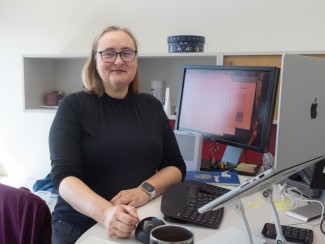 Woman with blonde hair and glasses sits at a desk with a computer monitor. She is turned to her right to face the camera