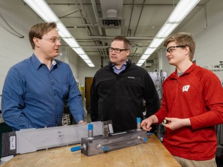 Three researchers wearing safety glasses discuss compact heat exchanger prototypes on a lab workbench, with a University of Wisconsin student in a red "W" pullover and two colleagues examining the metal test hardware.
