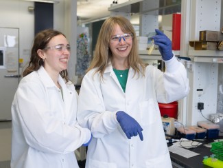A woman in a white lab coat and safety glasses holds up a flask of opaque yellow liquid as another woman in similar attire looks on smiling
