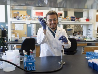 A smiling scientist in a white lab coat and purple nitrile gloves works with a pipette while standing at a black lab bench surrounded by various laboratory equipment including test tube racks, bottles, and other research instruments. 