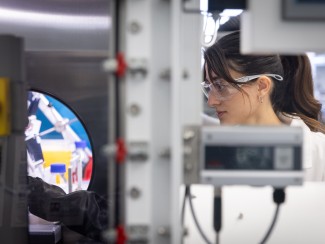 A researcher in a white lab coat and safety goggles looks through a circular portal into an automated laboratory workstation filled with robotic equipment and chemical vials.