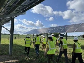 A group of visitors in high-visibility vests and hard hats tours an agrivoltaic solar installation, walking beneath elevated solar panels above a vegetated field on a partly cloudy day.
