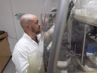 A bearded man in a white lab coat reaches for an object as he stands in front of a clear plastic barrier