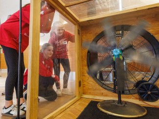 Three girls wearing matching red "Wind Storm" hoodies look through a clear panel into a wooden wind tunnel, watching a large fan spin rapidly.