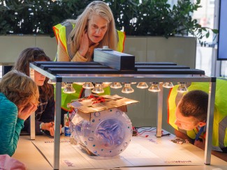 People stand around a table looking at a clear plastic cylinder with solar panels attached sitting under a bank of bright lights 