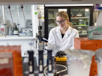 A woman in a white lab coat and lab glasses holds a pipette