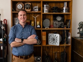 Dan Ludois stands in front of a brown bookcase filled with sundry items including books and antique electrical equipment