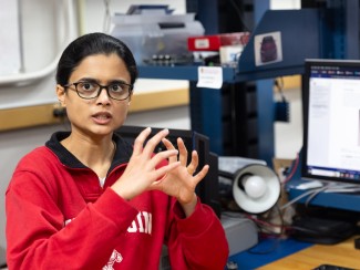 Woman with black hair and glasses in a red shirt gestures with her hands in front of a workbench with computer monitors and electronic equipment