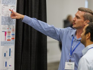 Man in blue and white shirt points to text on a poster as another man looks on