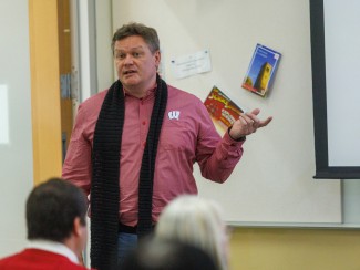 Man in red shirt with black scarf gestures as he talks to a room full of people