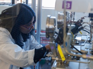 A researcher in a lab coat, safety glasses, and nitrile gloves sets up a high-pressure reactor system in a glass enclosure.