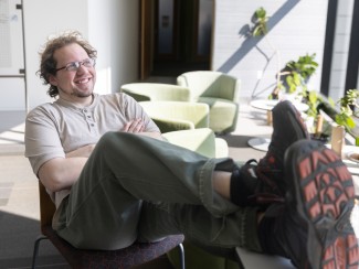A man reclines in a chair with his feet on a table in a sunlit space with potted plants