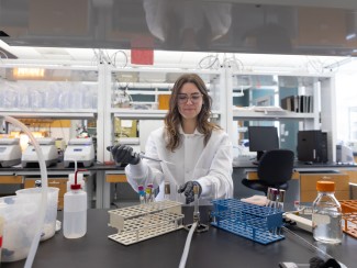 A woman in a white lab coat and latex gloves holds a flask of clear liquid in a laboratory