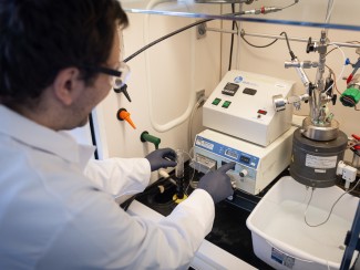 Man in white lab coat reaches into an exhaust hood to adjust equipment