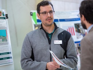 Man with glasses in gray pullover standing in front of an academic poster talking to another man whose back is to the camera
