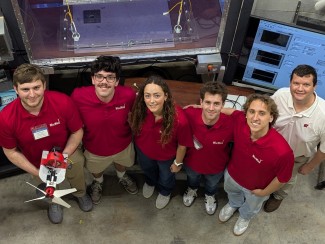 The WiscWind student team in matching red polos poses in front of a wind tunnel and data acquisition monitors, with one member holding their small-scale wind turbine prototype.