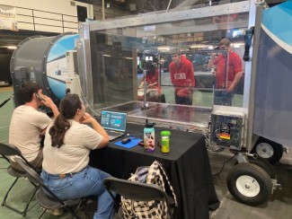 Two UW-Madison students sit at a control station with laptops, observing a model wind turbine being tested inside a clear-walled wind tunnel. Three people in red Wisconsin shirts work behind the transparent enclosure