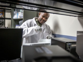 Man in white lab coat framed by dark shelving holds a glass vial