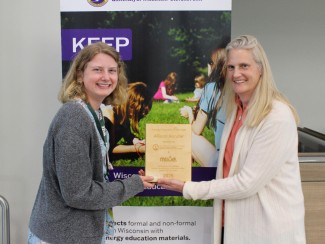 Two women smiling and holding a wooden plaque stand in front of a poster labeled KEEP with a photo of children on green grass