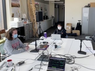 Mary Riker, left, sits at a table in front of a microphone. Michelle Chung, center, takes notes while Yiying Xiong, right answers a question on another microphone.