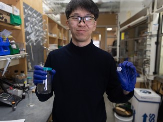 Smiling man standing in a laboratory holding a jar of black powder in one hand and a silver coin battery in the other