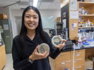 woman with long black hair standing in a lab holding two petri dishes