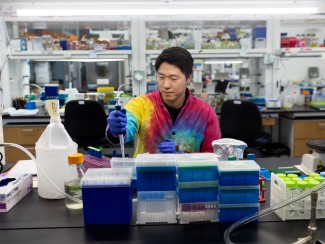 Asian man in tie-died lab coat seated at a lab bench