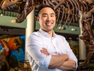 A smiling man with short dark hair, wearing a white button-down shirt with sleeves rolled up, stands with arms crossed in front of a large dinosaur skeleton display in a museum exhibit hall.